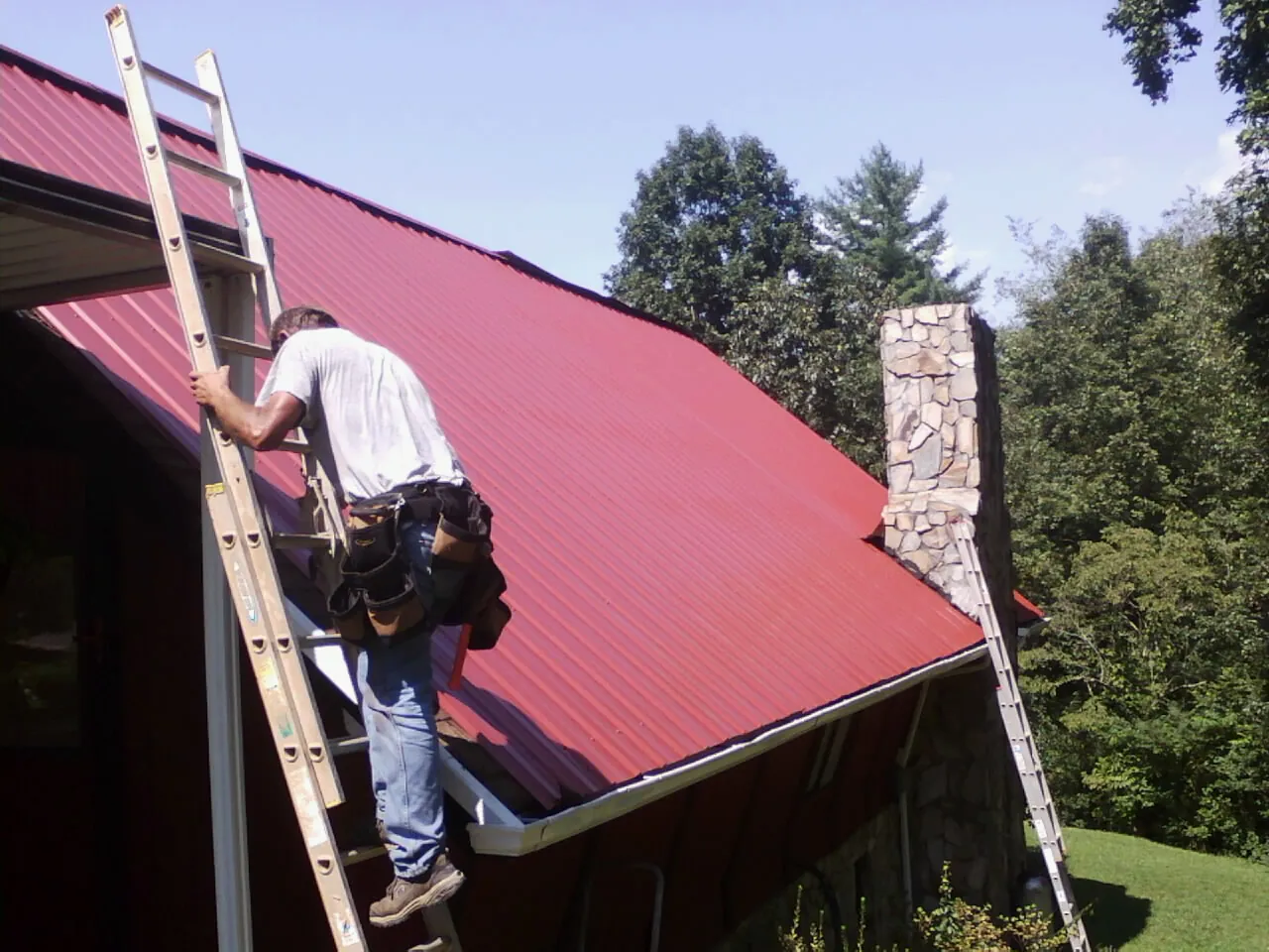 Roofer on a ladder beside a red metal roof and stone chimney — Soffit & Fascia Repair by Roofing Brothers in Wurtsboro